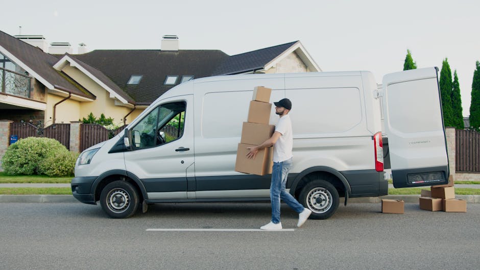 A man dressed in a white t-shirt, blue jeans, and a black cap stands inside the open rear of a white van parked on a paved surface, with his arms crossed. The van's interior is visible, showing a load
