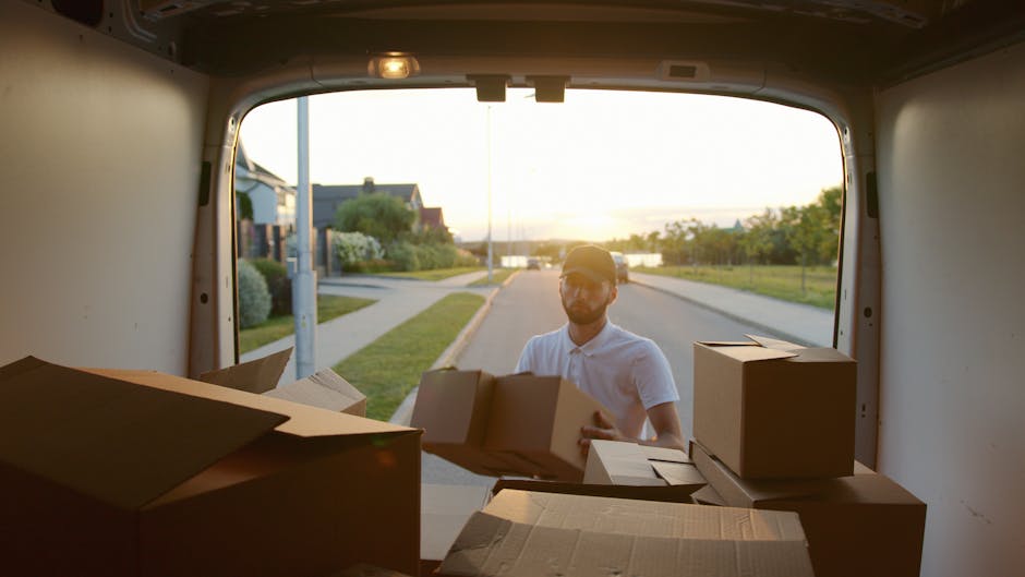 A man wearing a white polo shirt and sunglasses is seen from inside the back of a van, which is parked on a residential street during sunset. The van's open rear doors reveal various packed cardboard 