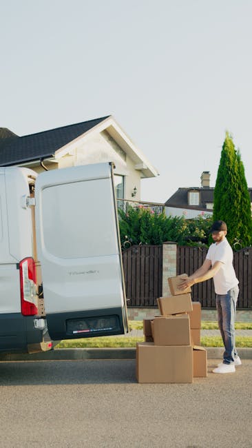 A man wearing a white polo shirt and sunglasses is seen from inside the back of a van, which is parked on a residential street during sunset. The van's open rear doors reveal various packed cardboard 