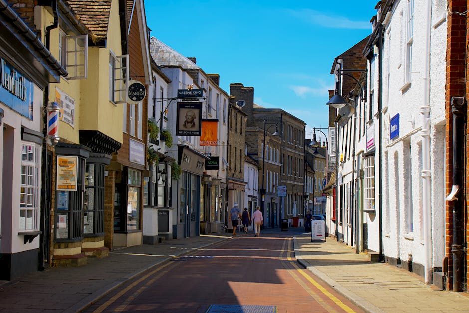 A narrow high street in Welling with colourful shopfronts, some with window displays and signage, and pedestrians walking along the sidewalk under clear blue skies. The buildings are a mix of traditional brick and painted facades, with adjacent doors and small awnings. The street surface is paved with bricks and features double yellow lines along the edge. In the foreground, the street is partly shaded by nearby buildings, while further down, the area is sunlit. This urban scene depicts a typical town centre environment suitable for house removals or furniture transport services. Man and Van Welling, a professional removals company, may be involved in home relocation or moving logistics in such environments, facilitating the loading and unloading of household goods in tight access areas, with vehicles parked close to the shopfronts and pedestrians present. The overall setting captures the essence of a bustling town street prepared for professional packing and moving operations.