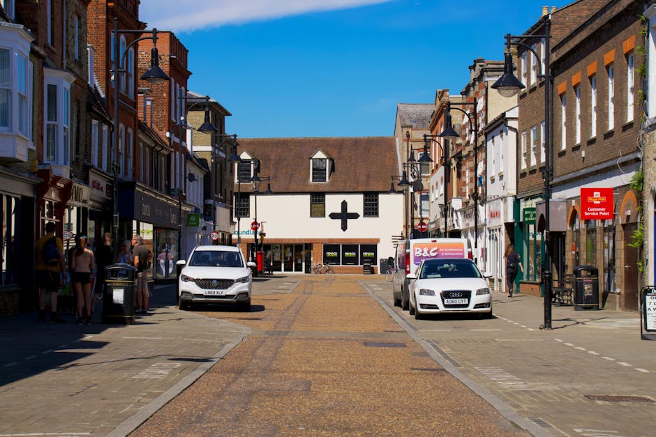 A street scene on Welling High Street showing a row of multi-storey brick and stucco commercial buildings with shops on the ground floor and residential entrances above, under a clear blue sky. The street pavement features a central textured, brownish strip, with parked vehicles including a white van, an Audi, and a B&C delivery truck positioned along the curbside. Pedestrians, some wearing casual clothing and hats, walk on the pavement and gather near the shop doorways. Streetlamps with curved arms and black finishes line both sides of the street, and a red customer service point sign is visible on one building. The area appears to be a busy commercial location, with elements indicating ongoing home relocation or furniture transport activities, as part of the moving process organized by Man and Van Welling, which specializes in removals services for challenging access jobs such as tight streets or limited entry points.