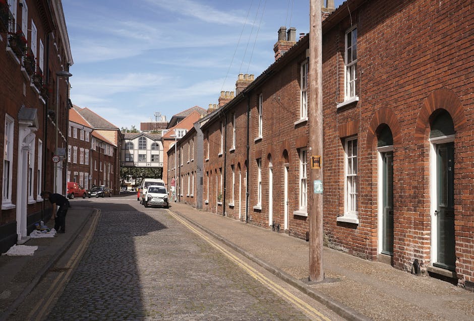 A narrow high street in Welling with colourful shopfronts, some with window displays and signage, and pedestrians walking along the sidewalk under clear blue skies. The buildings are a mix of traditional brick and painted facades, with adjacent doors and small awnings. The street surface is paved with bricks and features double yellow lines along the edge. In the foreground, the street is partly shaded by nearby buildings, while further down, the area is sunlit. This urban scene depicts a typical town centre environment suitable for house removals or furniture transport services. Man and Van Welling, a professional removals company, may be involved in home relocation or moving logistics in such environments, facilitating the loading and unloading of household goods in tight access areas, with vehicles parked close to the shopfronts and pedestrians present. The overall setting captures the essence of a bustling town street prepared for professional packing and moving operations.